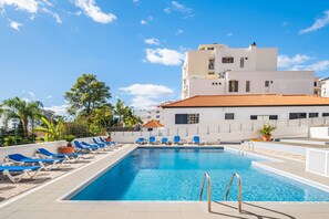 Una piscina con sillas reclinables azules y un edificio con techo de tejas al fondo.