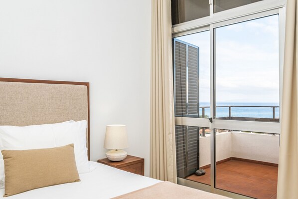 Habitación de hotel con cama, mesita de noche con lámpara y balcón con vista al mar.