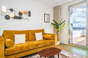A modern living room with a mustard yellow sofa, a wooden coffee table, a large potted plant, and a view of the balcony.