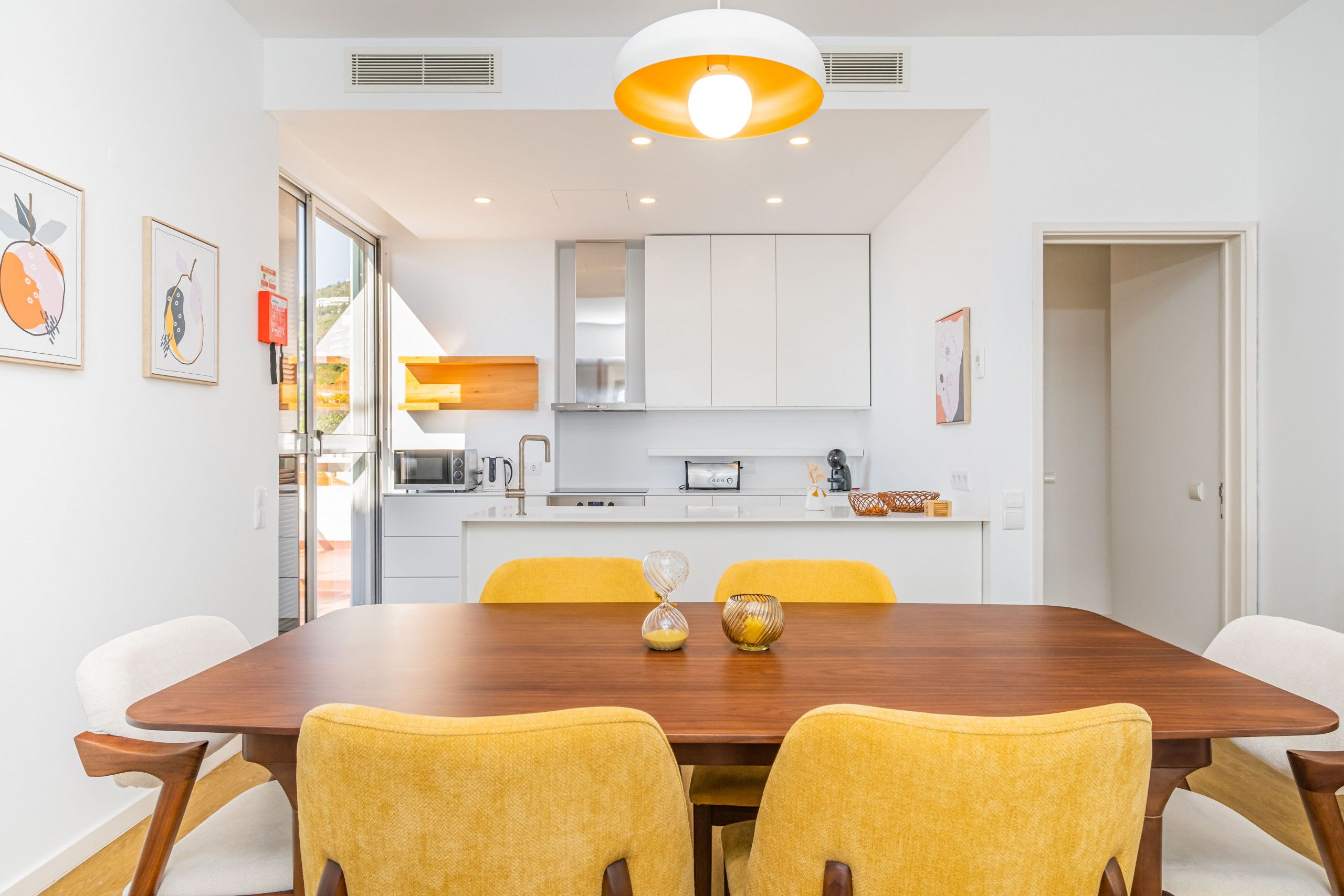 Un comedor moderno con una mesa de madera y sillas amarillas, paredes blancas y una cocina al fondo.