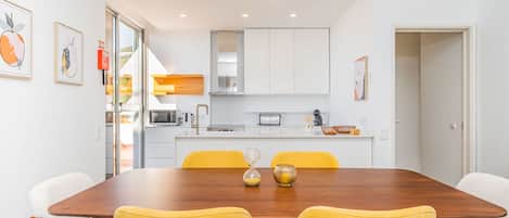 Un comedor moderno con una mesa de madera y sillas amarillas, paredes blancas y una cocina al fondo.