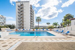 A swimming pool with blue lounge chairs, a tall building, and palm trees.