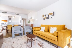 Una sala de estar moderna con un sofá amarillo, dos sillones y una mesa de centro. Se ve una cocina al fondo.