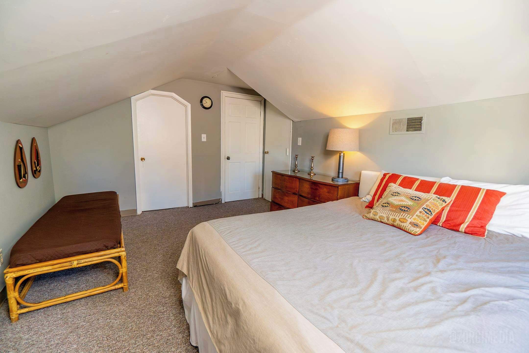 Un dormitorio con cama, un banco, cómoda, lámpara y un reloj en la pared.