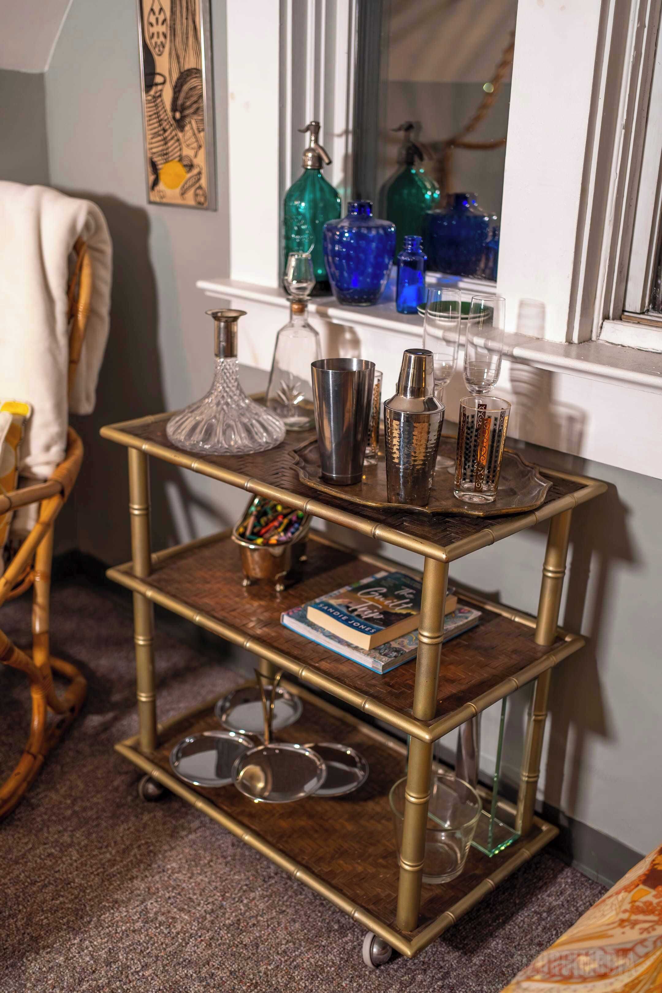 A wooden, three-tiered cart with various glassware and a book, placed in a room with a window and a framed picture on the wall.