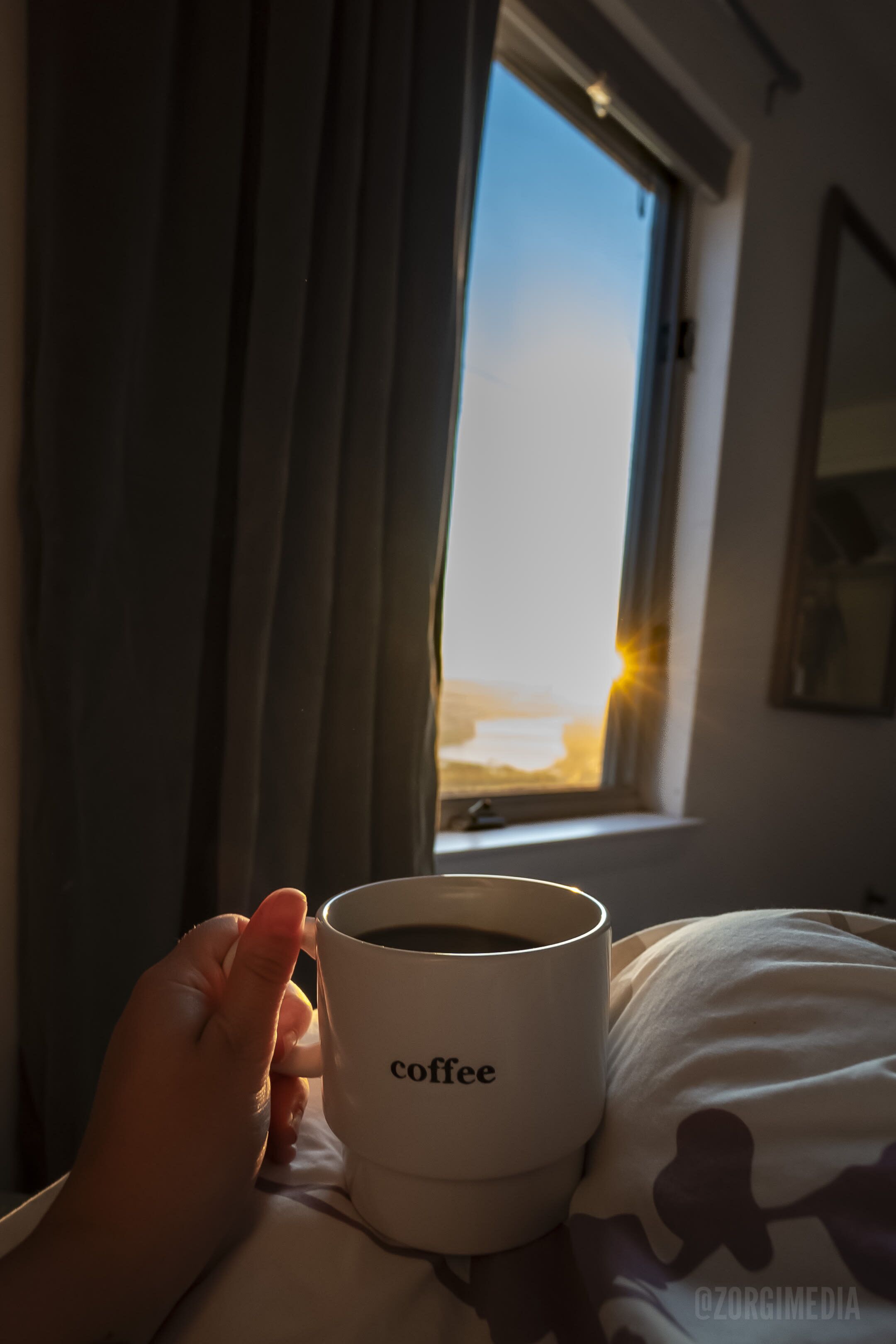 A hand holding a white mug labeled "coffee" on a bed with a view of a beach through a window.