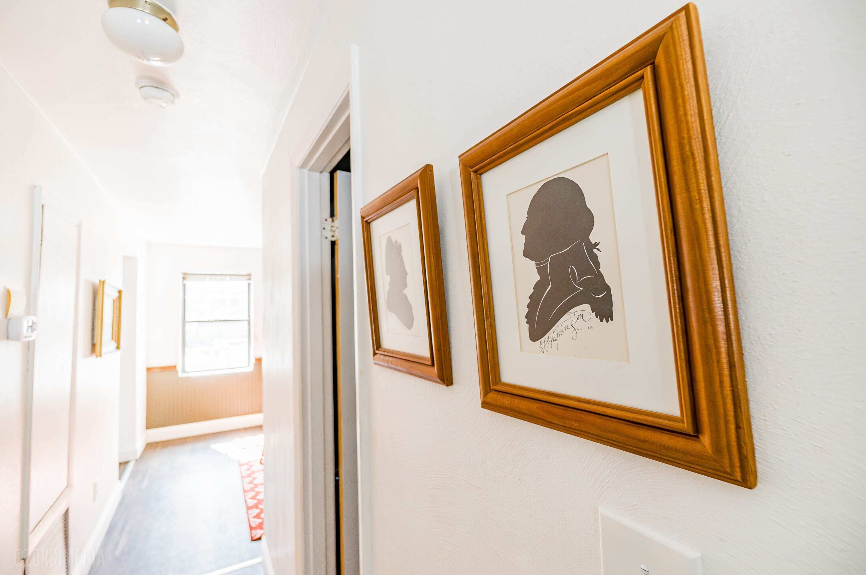 Un couloir aux murs ornés de tableaux encadrés, une porte donnant sur une autre pièce et un tapis rouge sur le sol.