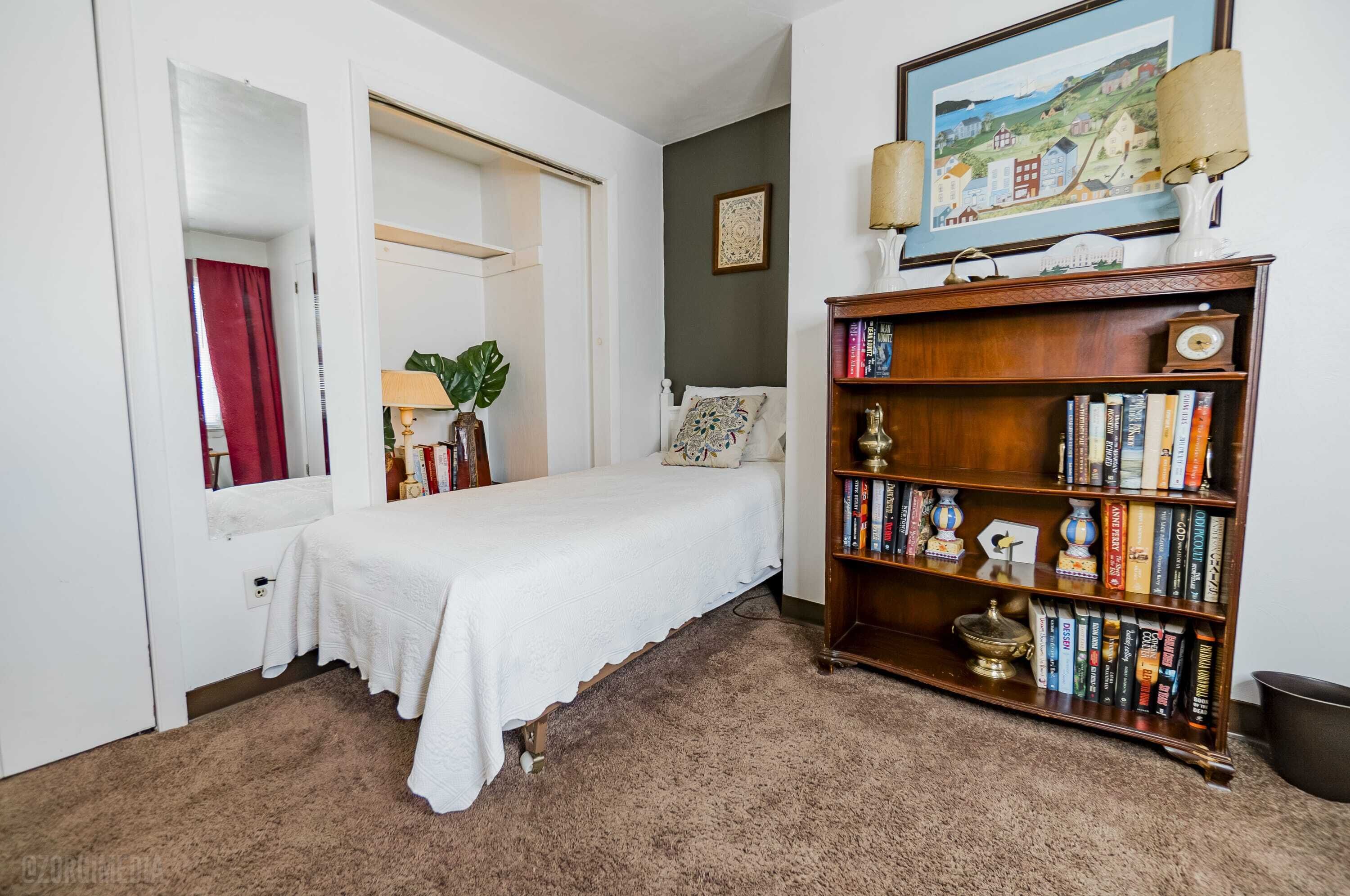 A bedroom with a bed, a bookshelf, a painting, and a lamp.