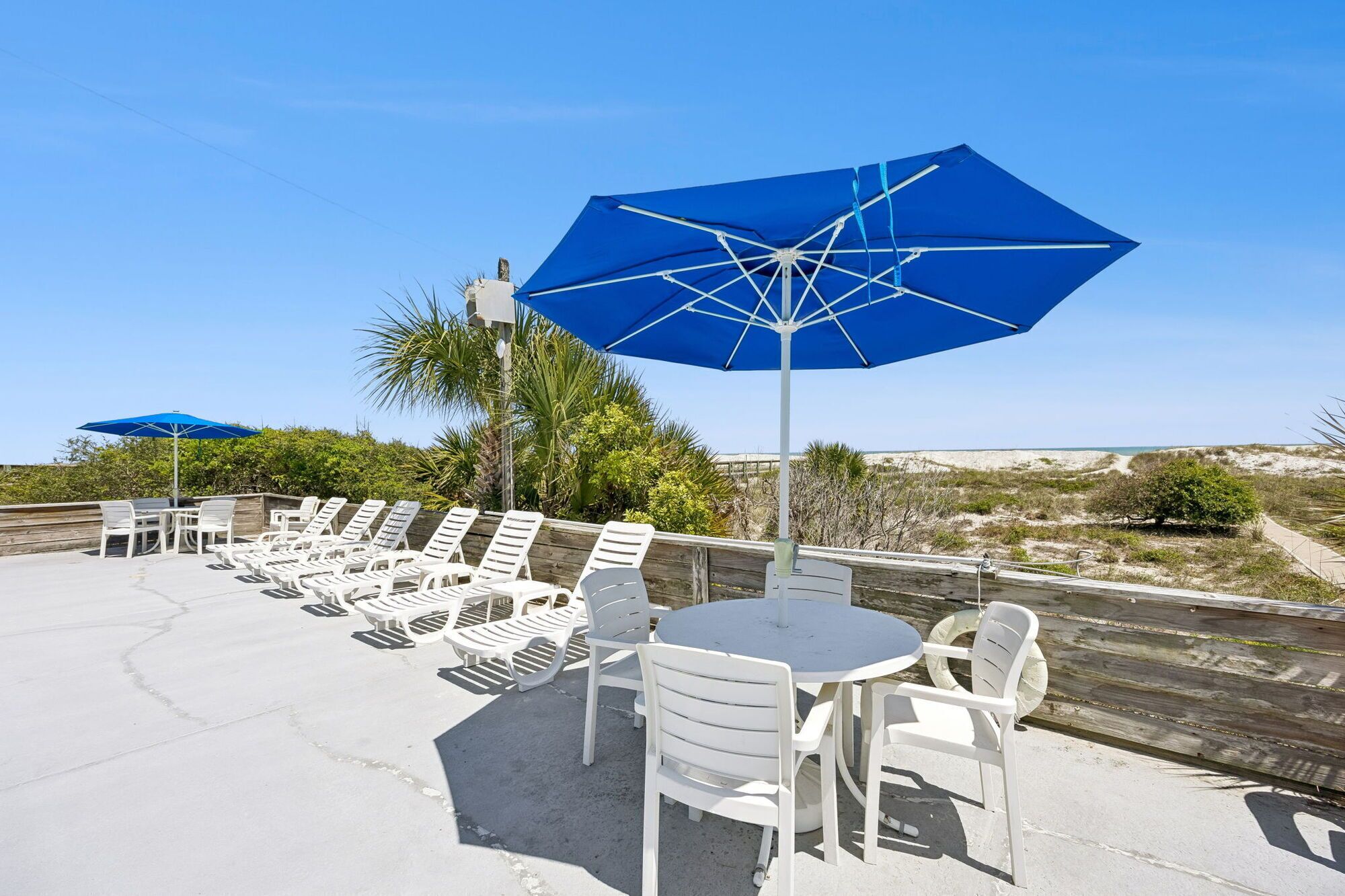 Une terrasse au bord de la plage, avec des chaises longues et des tables blanches, des parasols bleus et une vue sur la plage et les dunes.