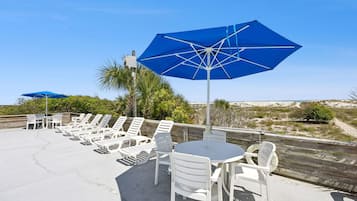 Une terrasse au bord de la plage, avec des chaises longues et des tables blanches, des parasols bleus et une vue sur la plage et les dunes.