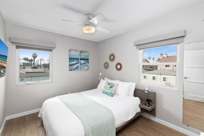A bedroom with a bed, a ceiling fan, two windows, and artwork on the walls.