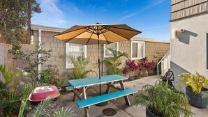 Une terrasse avec une table et un banc bleus, un parasol beige et divers plants en pot.