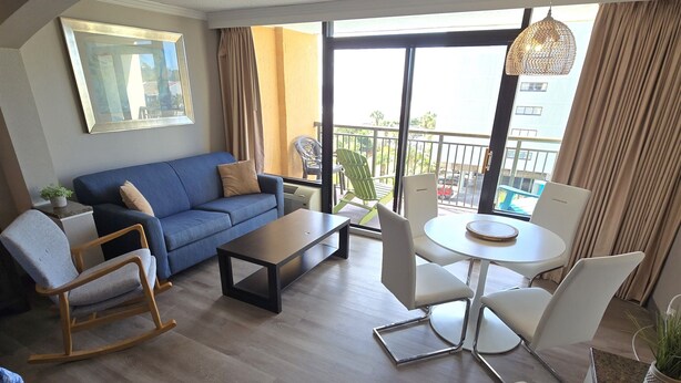 A modern living room with a blue sofa, a wooden rocking chair, a coffee table, and a dining table with white chairs. A large window offers a view of the outdoors. - Caravelle 505 Ocean view Efficiency w/pools (Myrtle Beach)