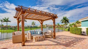 A covered outdoor seating area with wicker furniture and a pergola, overlooking a landscaped garden and a body of water.