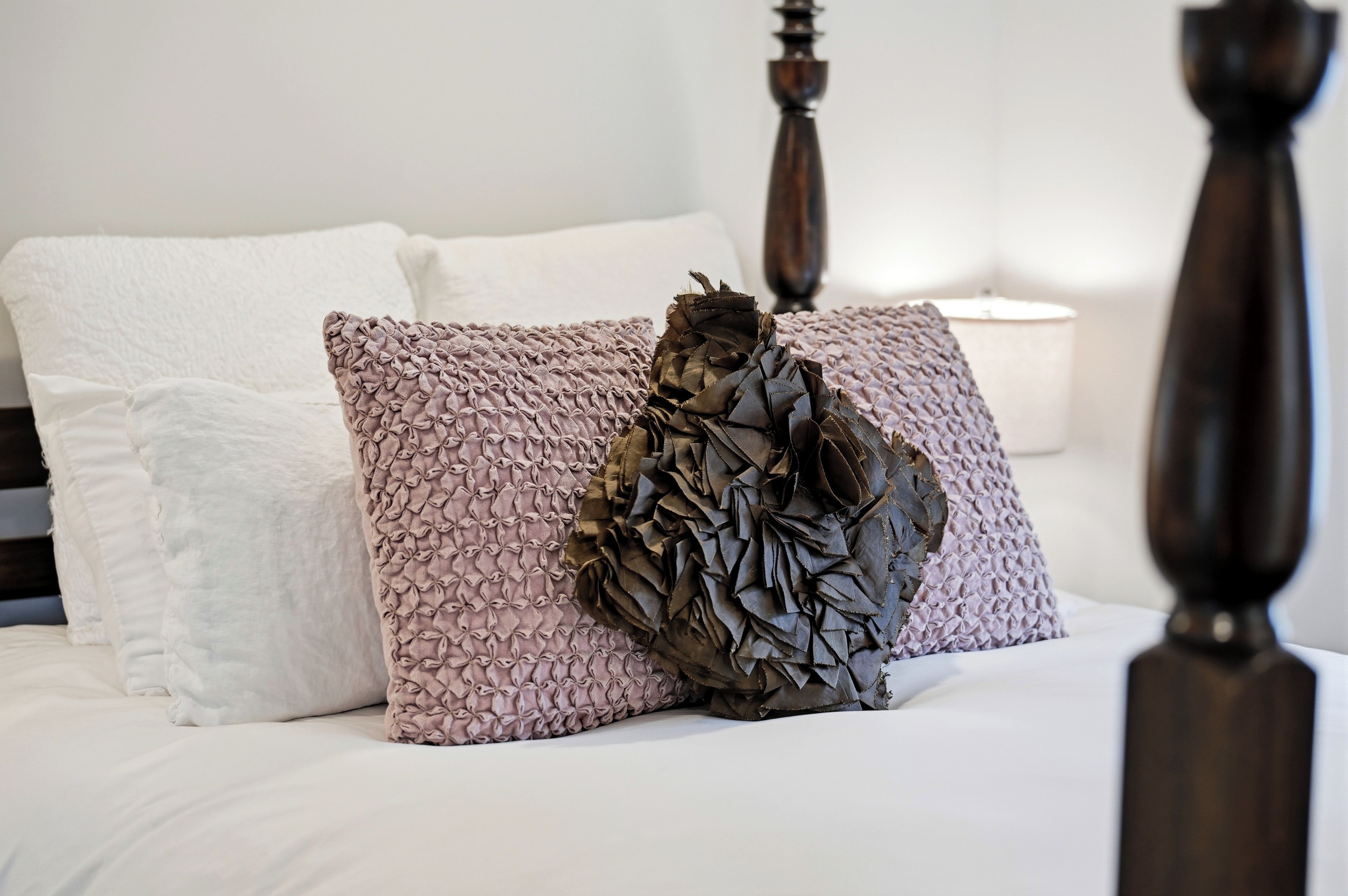 A bed with a white and pink textured pillow, a decorative leaf-shaped pillow, and a white bedside lamp.