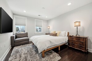 Une chambre à coucher comprenant un lit, une commode, un téléviseur et deux fenêtres équipées de stores.