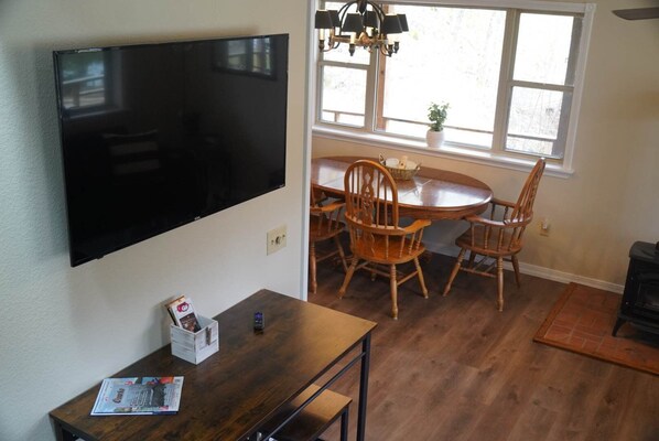 A dining area with a round table and four wooden chairs, a TV mounted on the wall, and a window with a view of the outdoors.