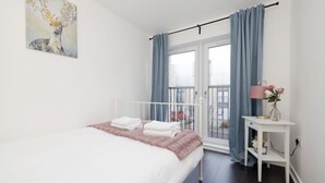 A bedroom with a bed, a bedside table with a lamp and flowers, a window with curtains, and a painting on the wall. - Stylish House with Garden & Parking (Glasgow)