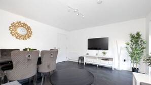 A modern living room with a round black table, grey upholstered chairs, a white console table with a flat-screen TV, and a decorative wall piece. - Stylish House with Garden & Parking (Glasgow)