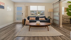 A living room with a dark blue sofa, two patterned pillows, a wooden coffee table, and a large window with blinds. - Family Beach Retreat | Pool + Golf Cart (Jacksonville Beach)