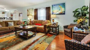 A spacious living room with a large sofa, a coffee table, and a patterned rug. There is a dining area in the background with a table and chairs.