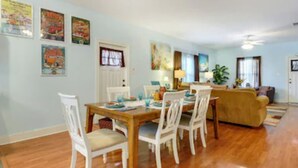 A dining area with a wooden table and white chairs, a living room with a sofa and a TV, and a kitchen in the background.