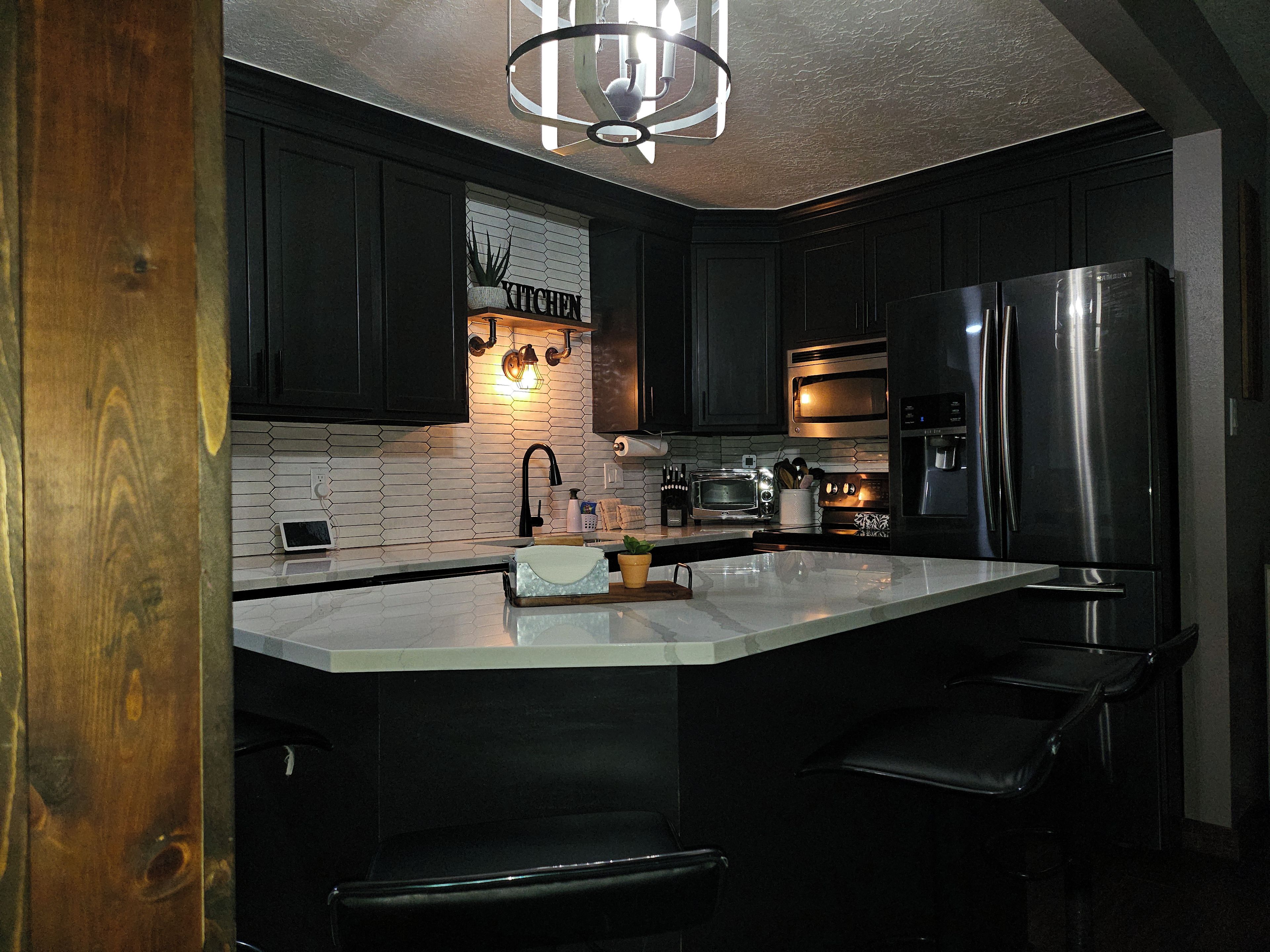 A modern kitchen with dark cabinetry, a white countertop, a built-in oven, a stainless steel refrigerator, and a pendant light above the island.