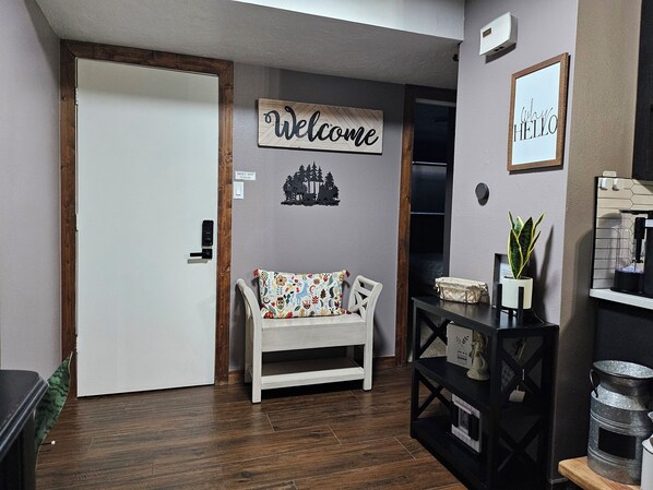 A hallway with a white door, a bench with a colorful cushion, a welcome sign, a small potted plant, and a metal container on a shelf.