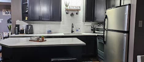 A modern kitchen with dark cabinets, a stainless steel refrigerator, a built-in oven, and a large island with a coffee maker and toaster.