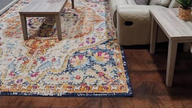 Ein Wohnzimmer mit einem grauen Ecksofa, einem kleinen Holztisch und einem farbenfrohen Teppich.