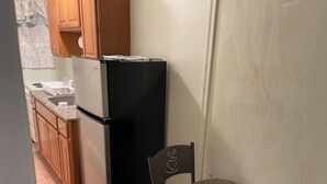 Une cuisine compacte avec un réfrigérateur noir, des placards en bois, une petite table à manger agrémentée d’une plante en pot et une chaise.