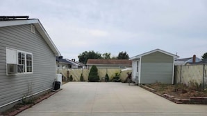 Un jardin à l’arrière d’une maison, avec une terrasse dallée, un petit abri de jardin et une zone délimitée par une clôture, où se trouvent des arbres.