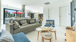 Un salón moderno con un sofá gris, un sillón azul y una mesa de centro de madera. Se ve una ventana grande con sombrillas rojas en el exterior.
