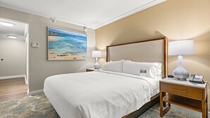 A hotel room with a large bed, a nightstand with a lamp, a painting of a beach scene, and a door leading to another room. - Maui Resort Rentals: Marriott's Maui Ocean Club Studio Oceanfront Suite (Lahaina)