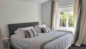 A neatly made bed with a grey headboard, white bedding, and striped pillows. There is a window with curtains, and a small bedside table with a lamp.