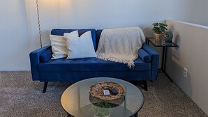 Un sofá azul con un plaid blanco, una mesita redonda con una cesta tejida y una lámpara de pie.
