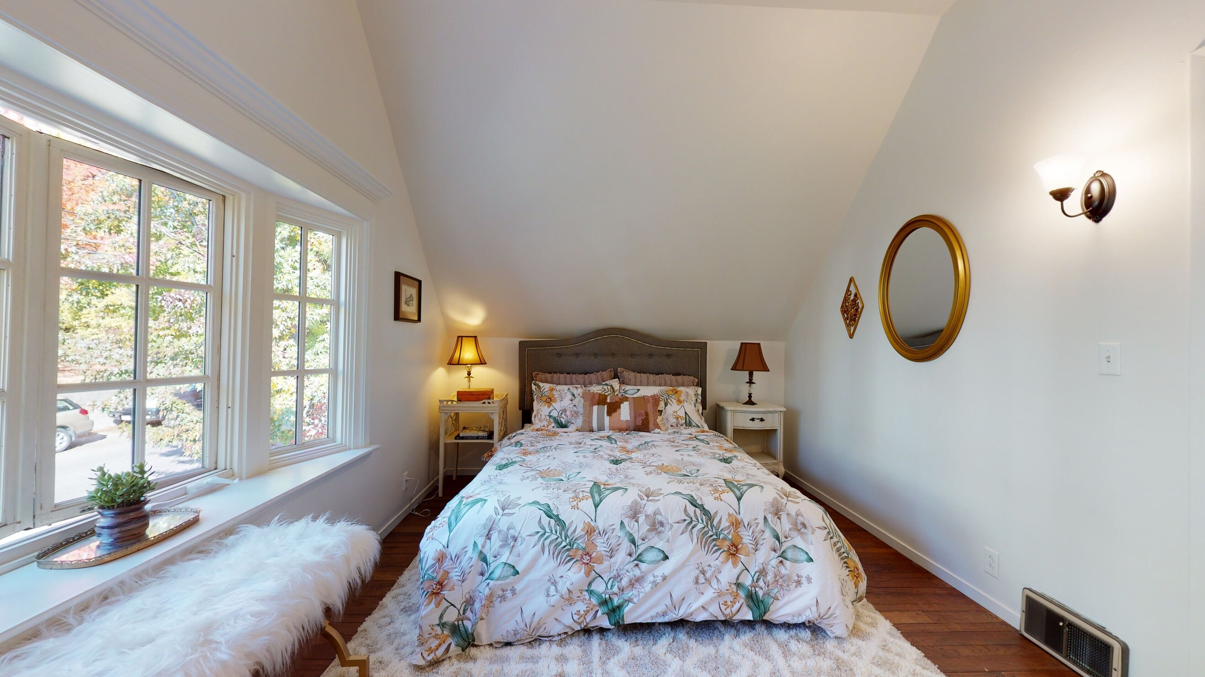 A bedroom with a large window, a bed with floral bedding, a nightstand, a lamp, a mirror, and a radiator.