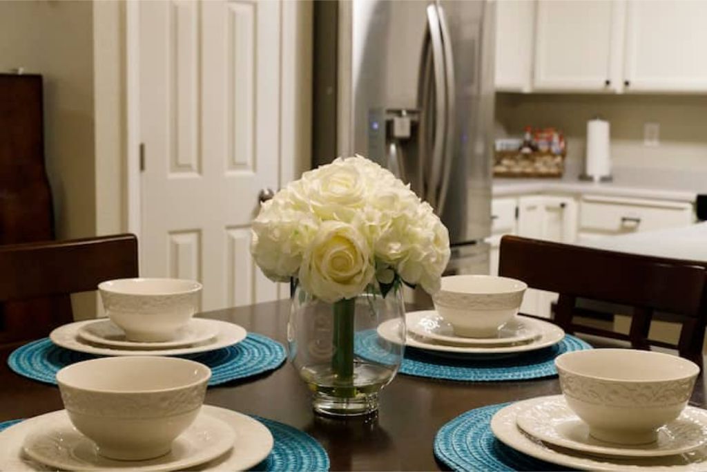 Une table à manger avec des bols blancs et des sets de table bleus, un vase avec des fleurs blanches et, au fond, un réfrigérateur en acier inoxydable.