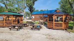 A rustic wooden cabin with a green roof, surrounded by picnic tables and benches, set in a gravel area with trees in the background. - The Getaway #15 (Ashtabula)