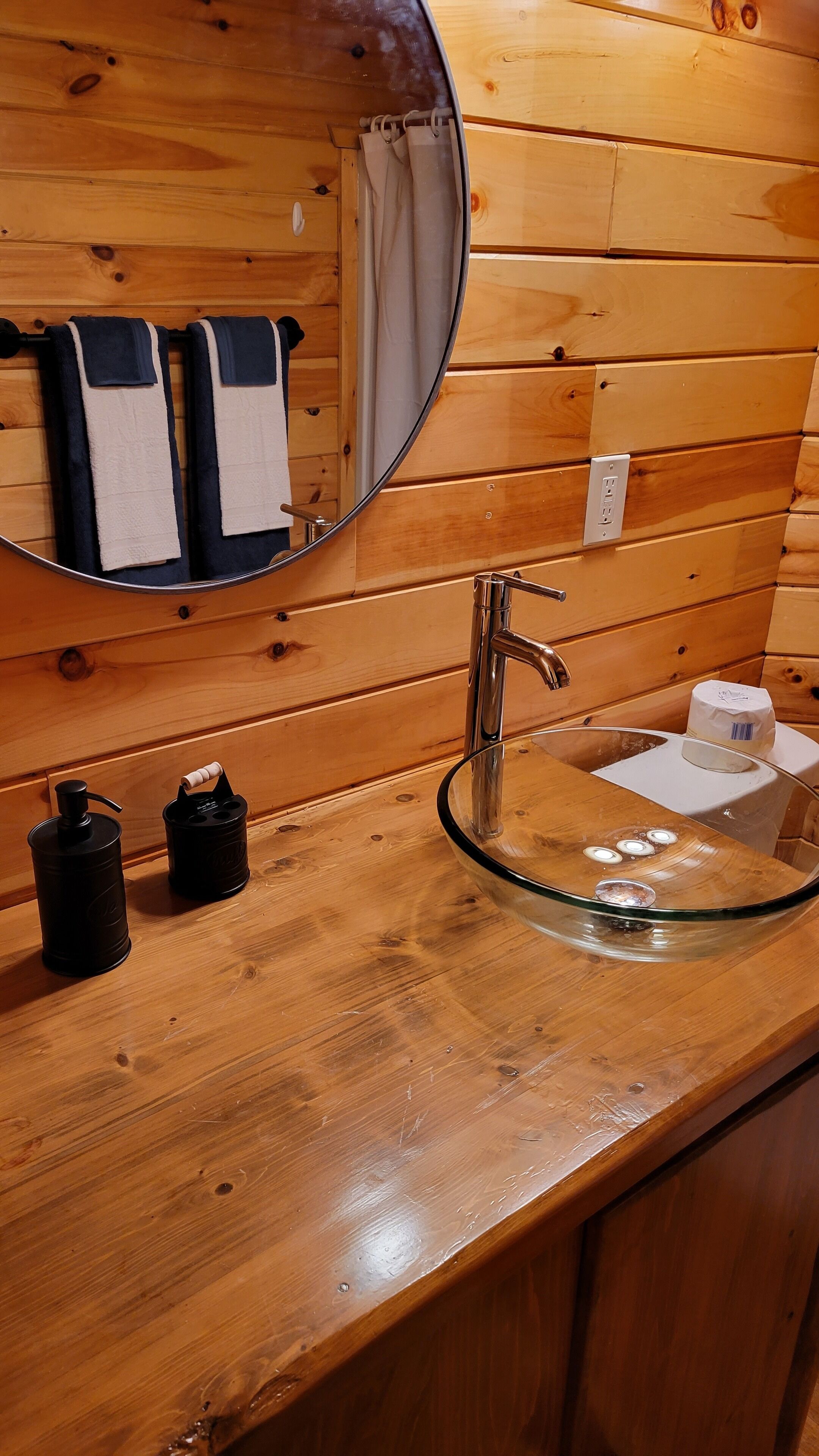 A wooden bathroom vanity with a glass sink, a mirror, and two black dispensers.