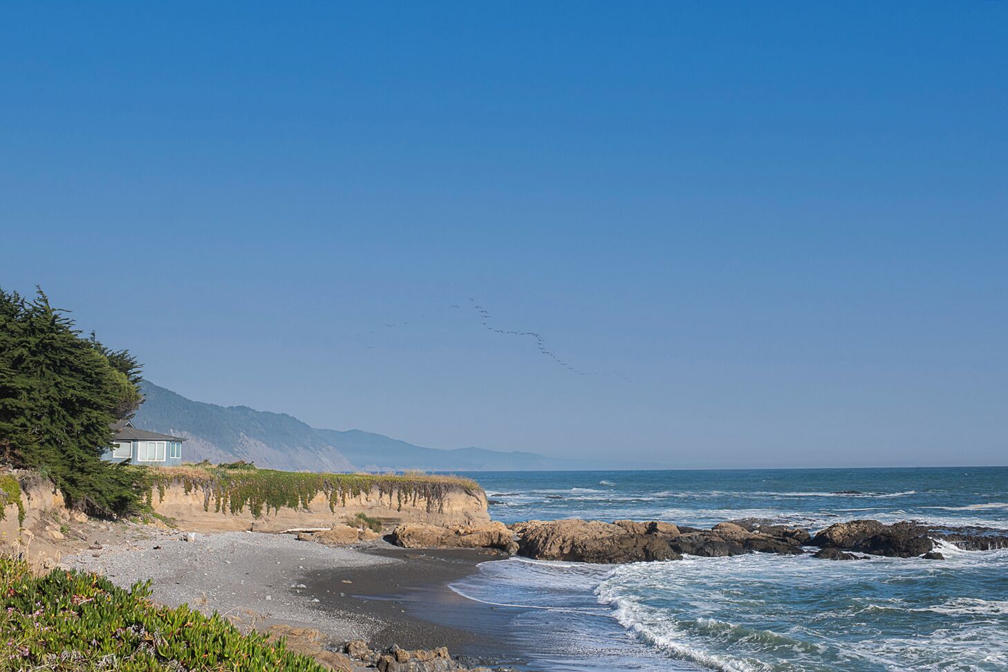 A coastal landscape with a house on a cliff, rocky shoreline, and waves crashing onto the beach.