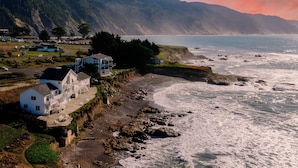 A coastal house with a balcony overlooking the sea, situated on a rocky shoreline with a view of the ocean and distant mountains. - Black Sands Inn (Shelter Cove)