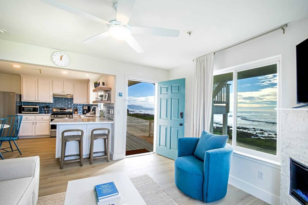 A modern kitchen with a central island, a dining area with blue chairs, a living room with a blue sofa, and a large window with a view of the ocean.