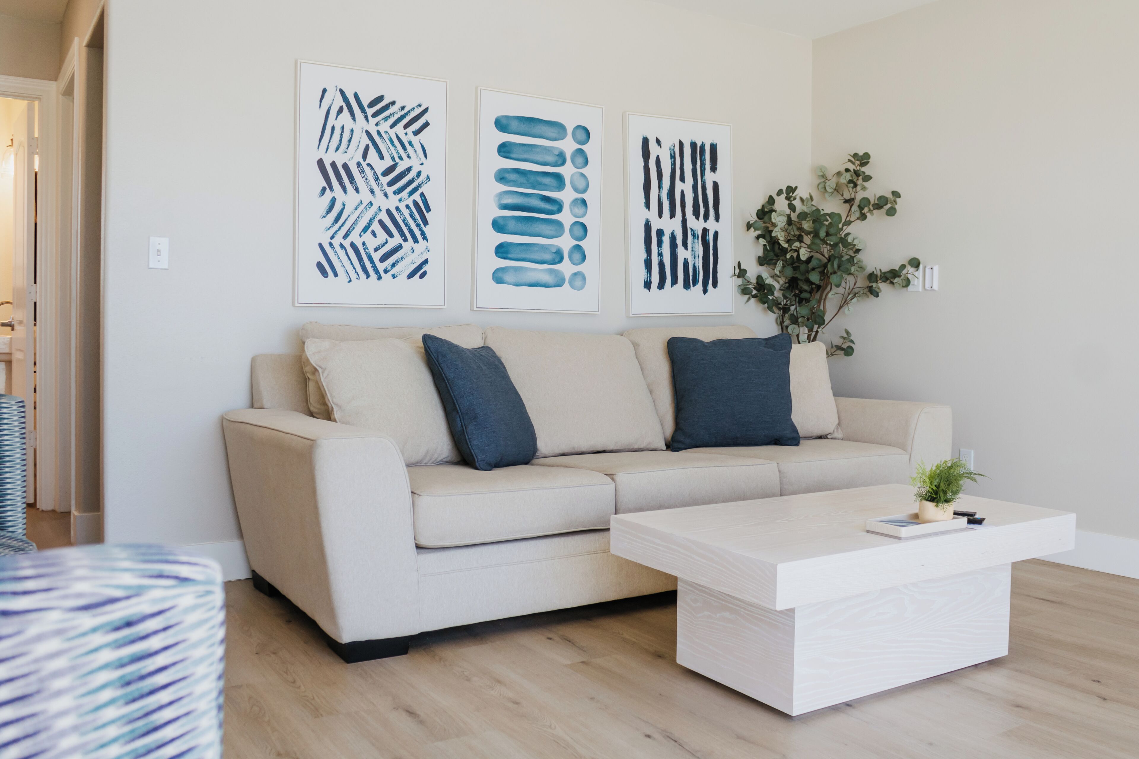 A modern living room with a beige sofa, two dark blue pillows, a white coffee table, and three abstract art pieces on the wall.