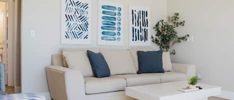 A modern living room with a beige sofa, two dark blue pillows, a white coffee table, and three abstract art pieces on the wall.