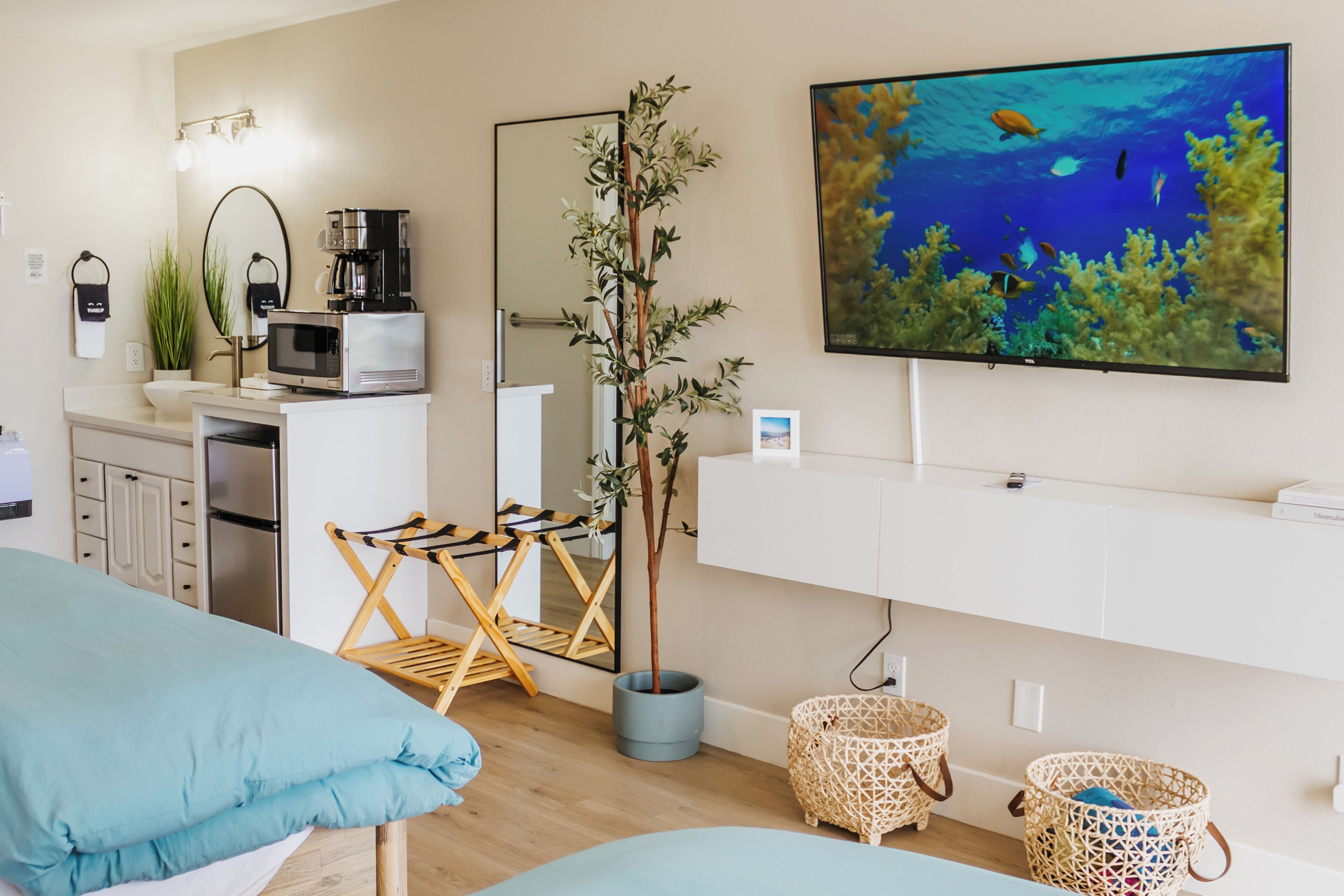 A modern room with a bed, a TV displaying an underwater scene, a microwave, a coffee maker, a plant, and a small wooden chair.