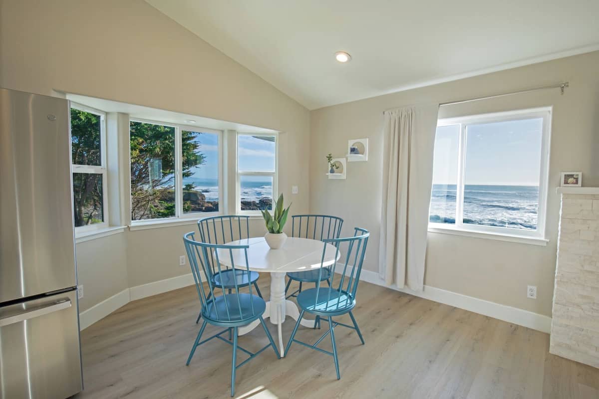 A dining area with a white table and blue chairs, large windows with ocean views, and a modern kitchen with a stainless steel refrigerator.