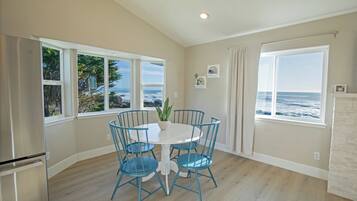 A dining area with a white table and blue chairs, large windows with ocean views, and a modern kitchen with a stainless steel refrigerator.