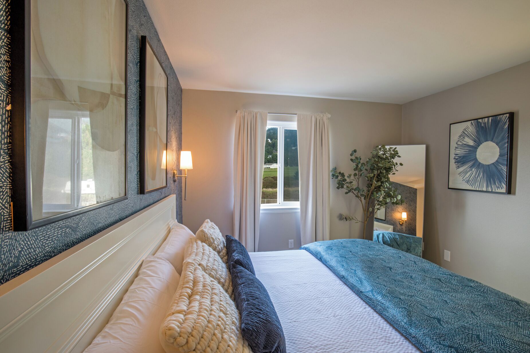 Una habitación de hotel con una cama grande, una ventana con cortinas, un espejo, una lámpara de pared y una obra de arte enmarcada.