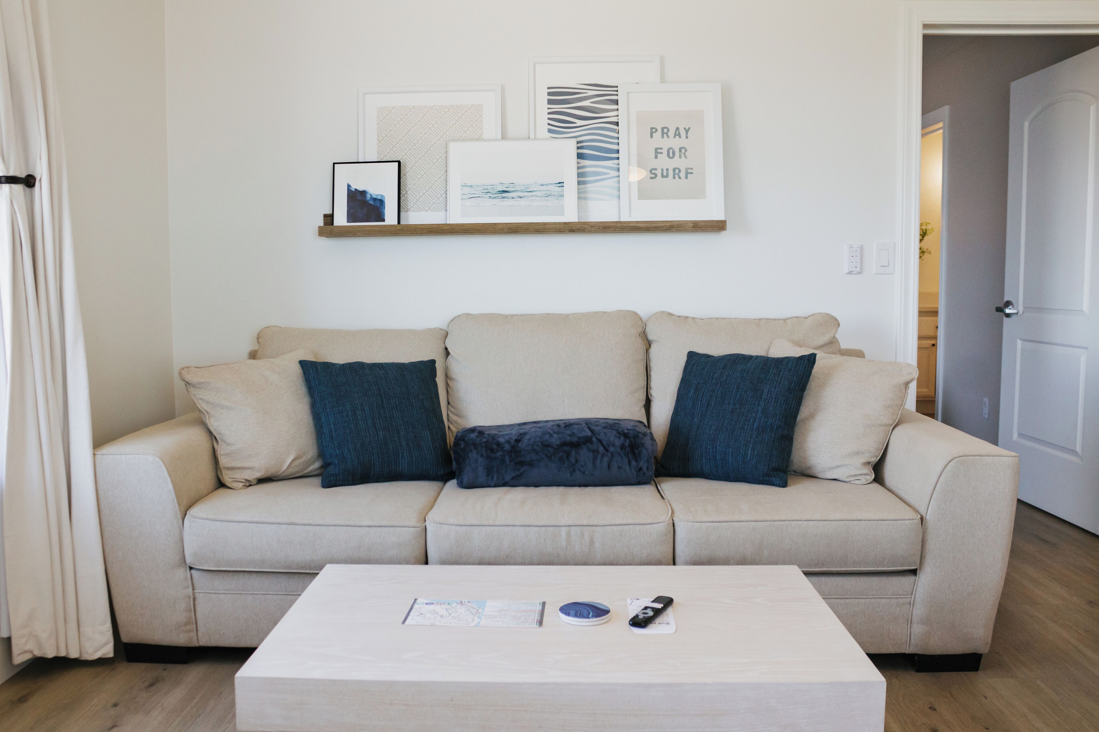 A beige sofa with three blue cushions, a white coffee table with a remote and a map, and a wall with framed artwork including a 'PRAY FOR SURF' sign.